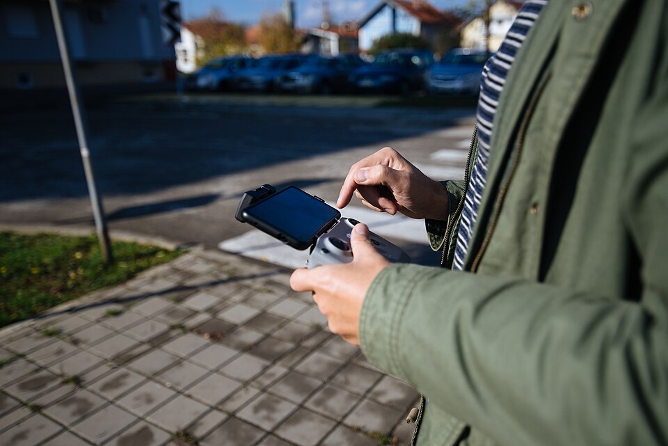 A person stands on a sidewalk, using a drone controller to navigate their device. Surrounding is a q