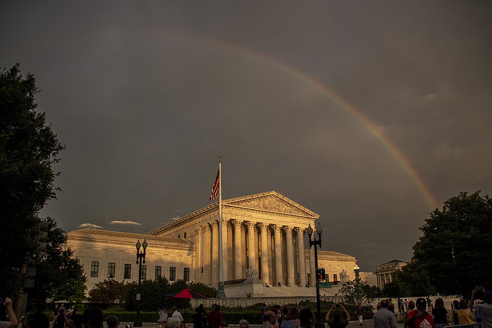 A colorful rainbow arches over a building with tall vertical columns holding up the peaked roof and 